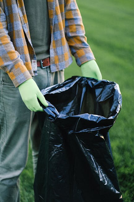 An expansive landfill site filled with layers of mixed waste, including plastic bags, discarded clothing, and household items, spread across uneven terrain with mounds of refuse. Several yellow Komats