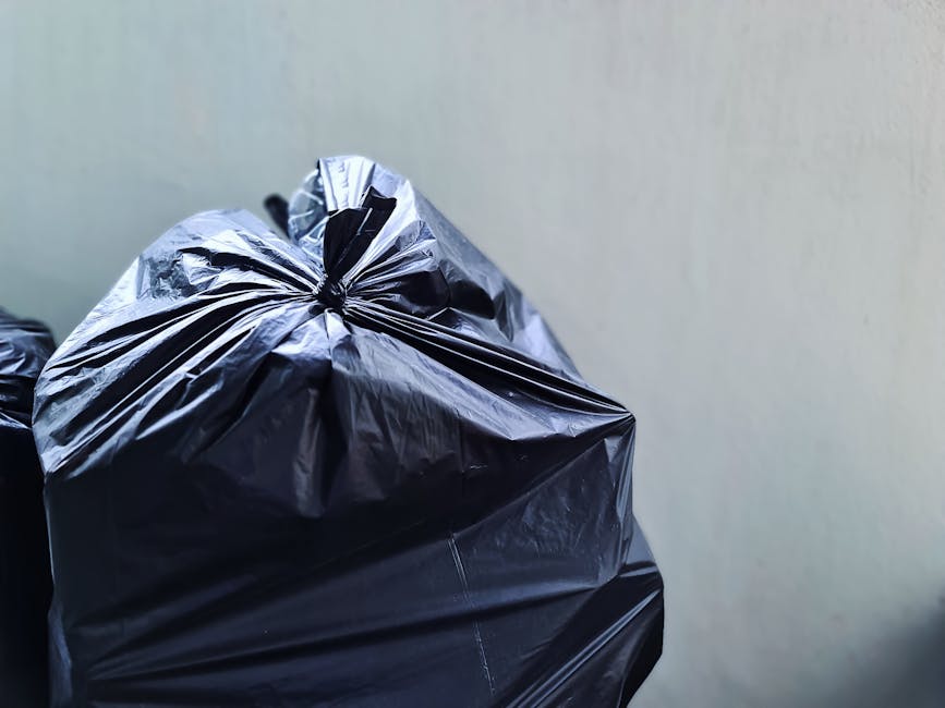 A green wheeled waste bin filled with multiple red and black bags containing garden or general waste, placed on a concrete pavement against a dark brick wall background. Adjacent to the bin, there is 